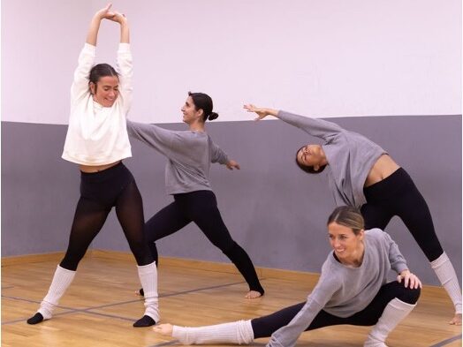 4 chicas bailando en un aula con suelo de madera frente a una pared blanca y gris.
