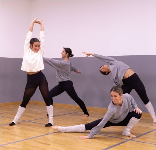 4 chicas bailando en un aula con suelo de madera frente a una pared blanca y gris.